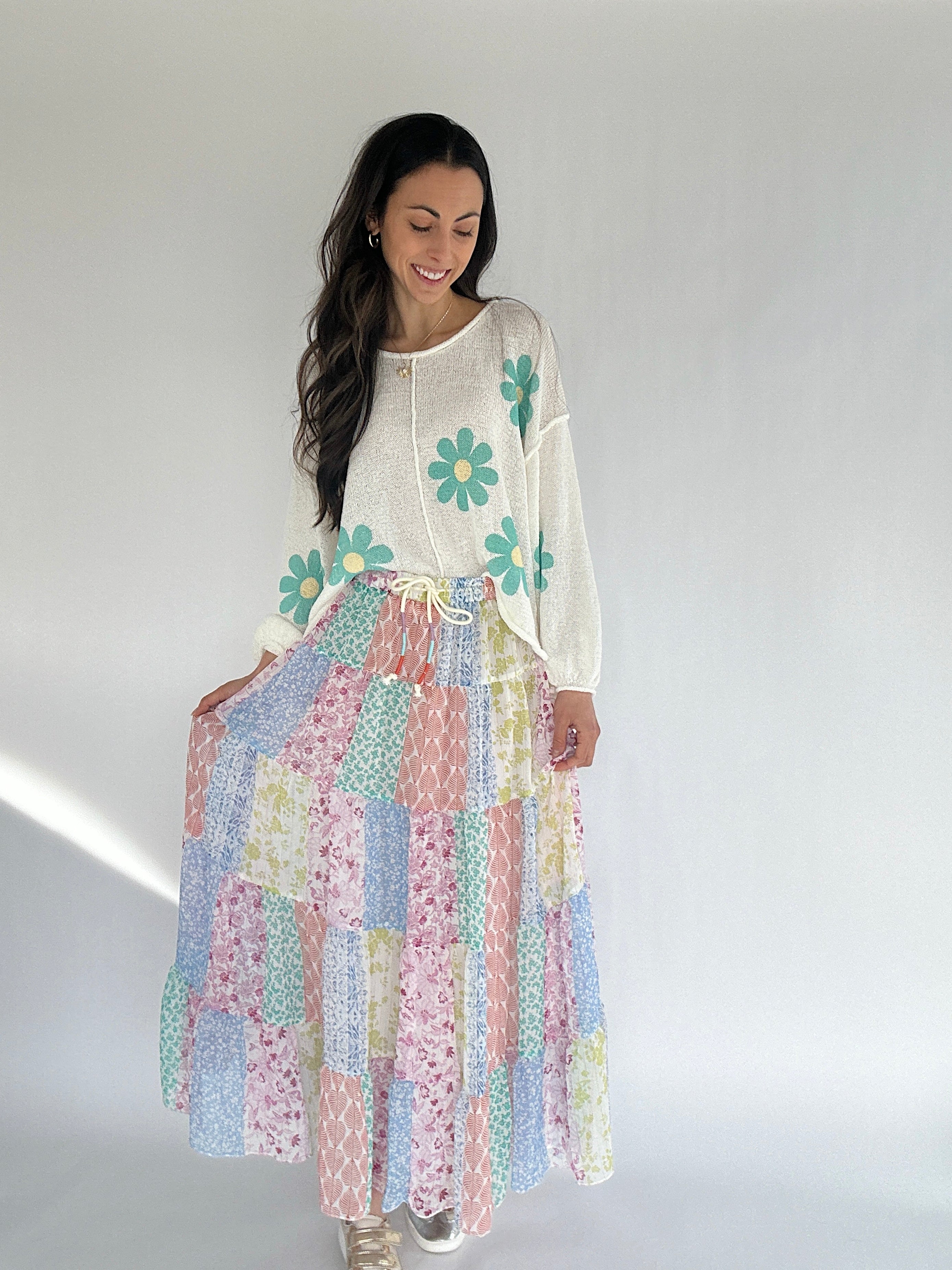 Woman wearing a long sleeve ivory sweater with daisies and colorful patchwork printed maxi skirt against a plain background