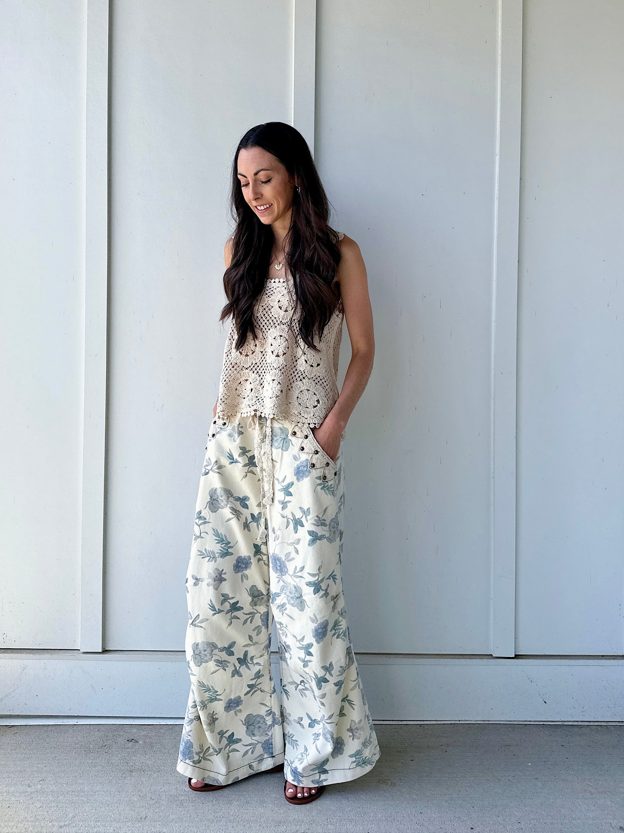 Woman wearing a crochet top and floral wide-leg pants with hands in pockets against a white wall.