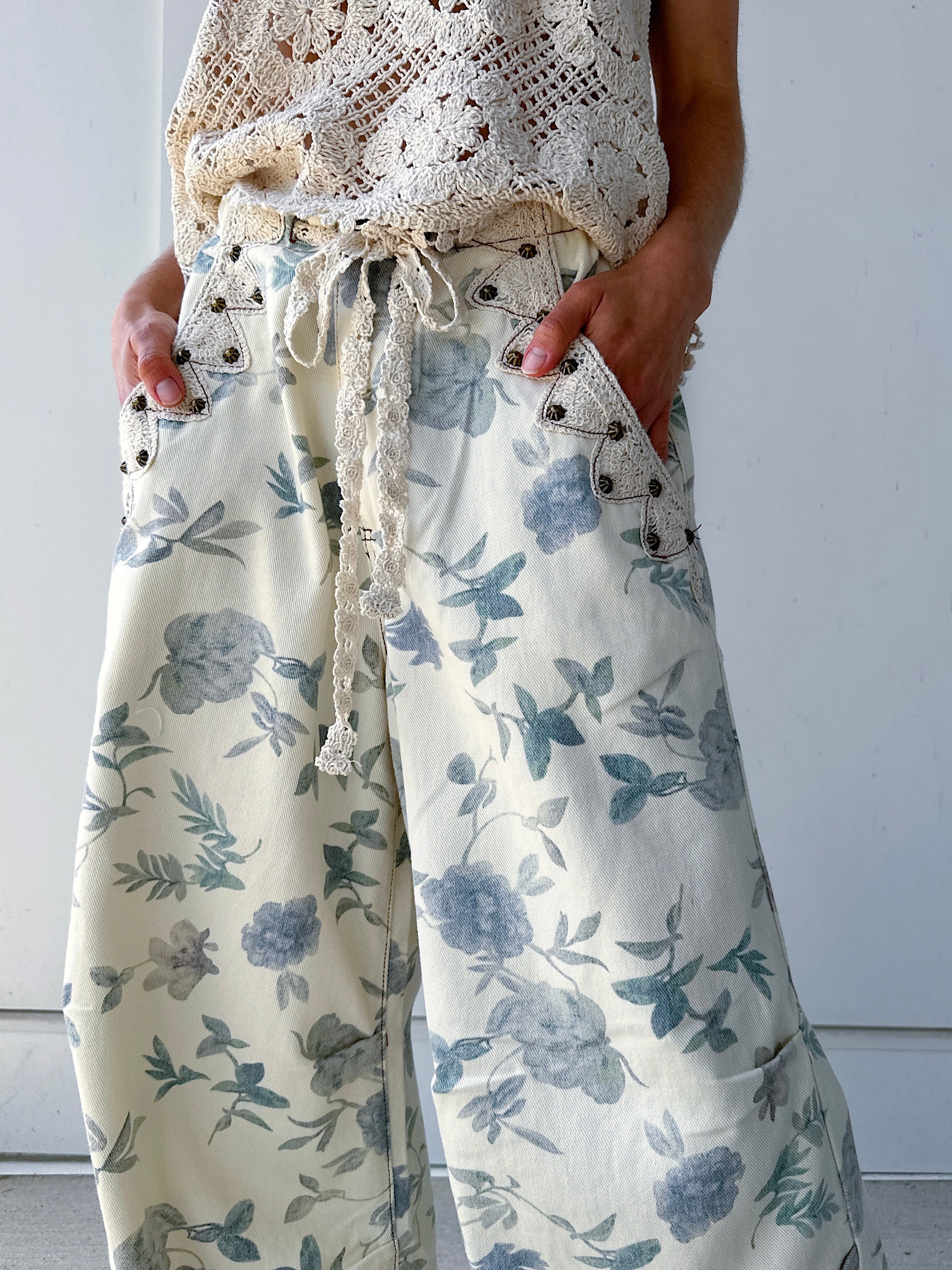 Person wearing a white lace top and floral pants against a plain background with hands in pockets that are studded and lined by crochet trim