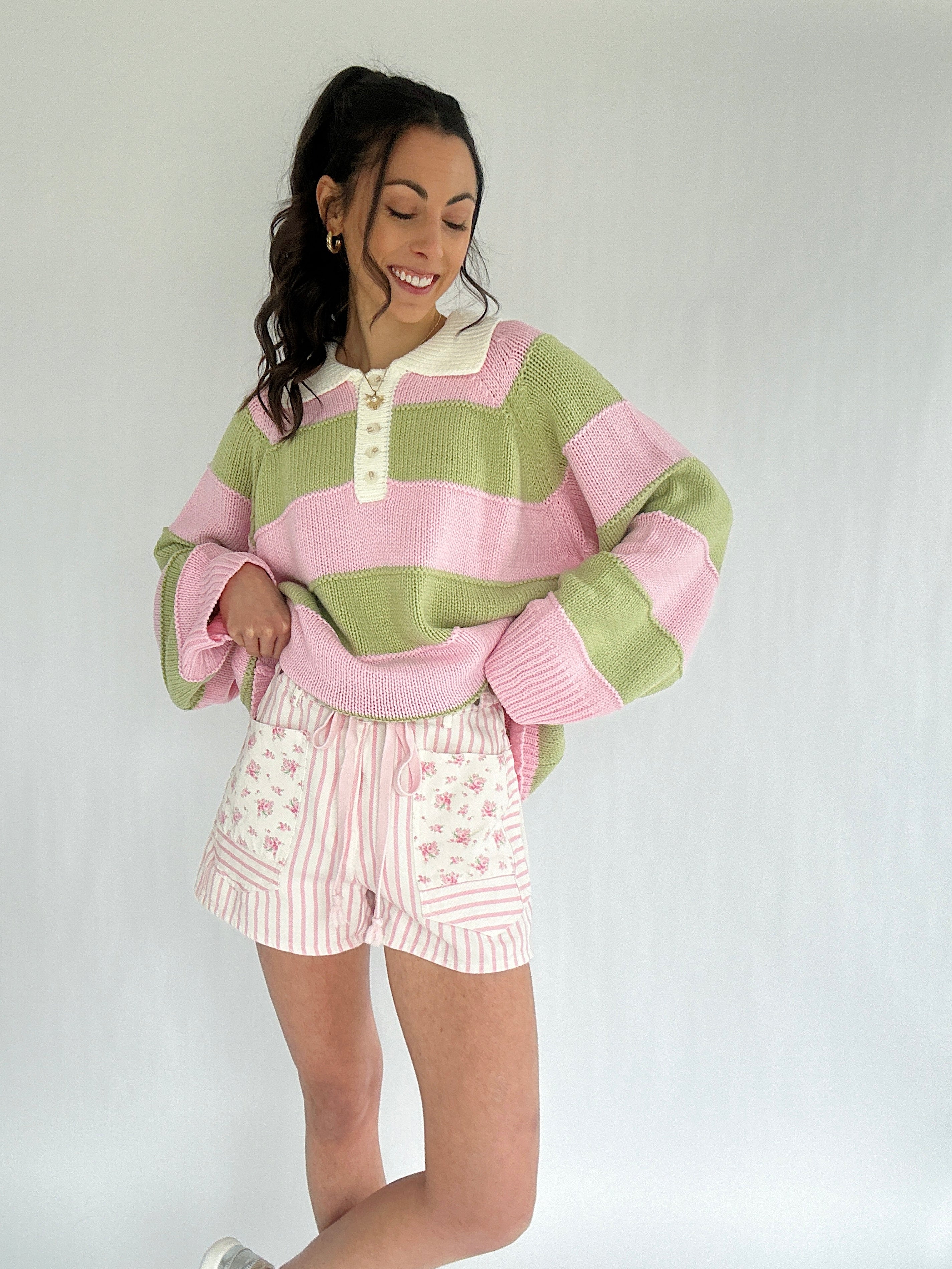 Woman wearing a green and pink striped sweater with white floral and striped shorts on a plain background
