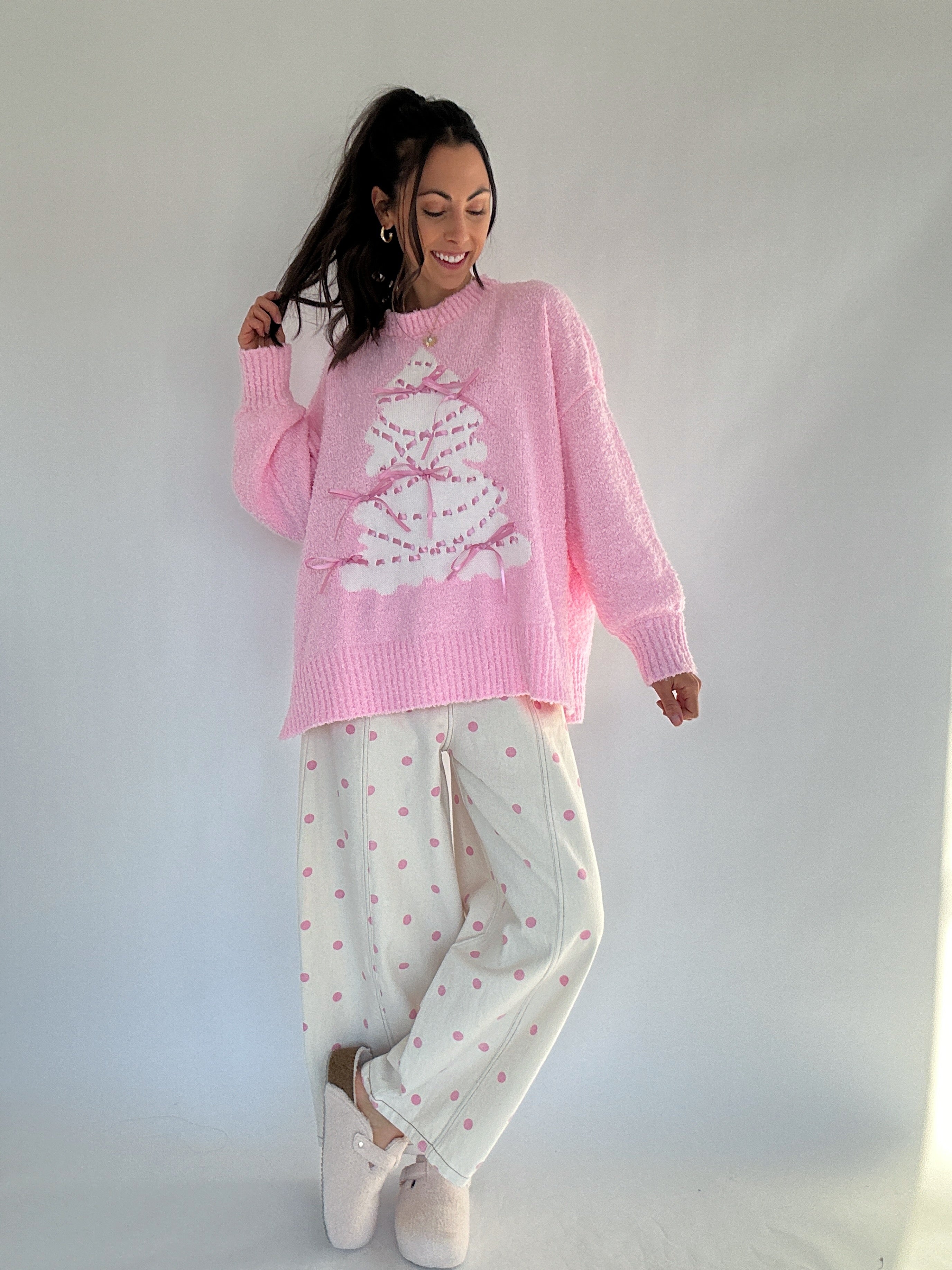 Woman wearing a pink sweater with a white tree design and pink polka dot pants against a white background