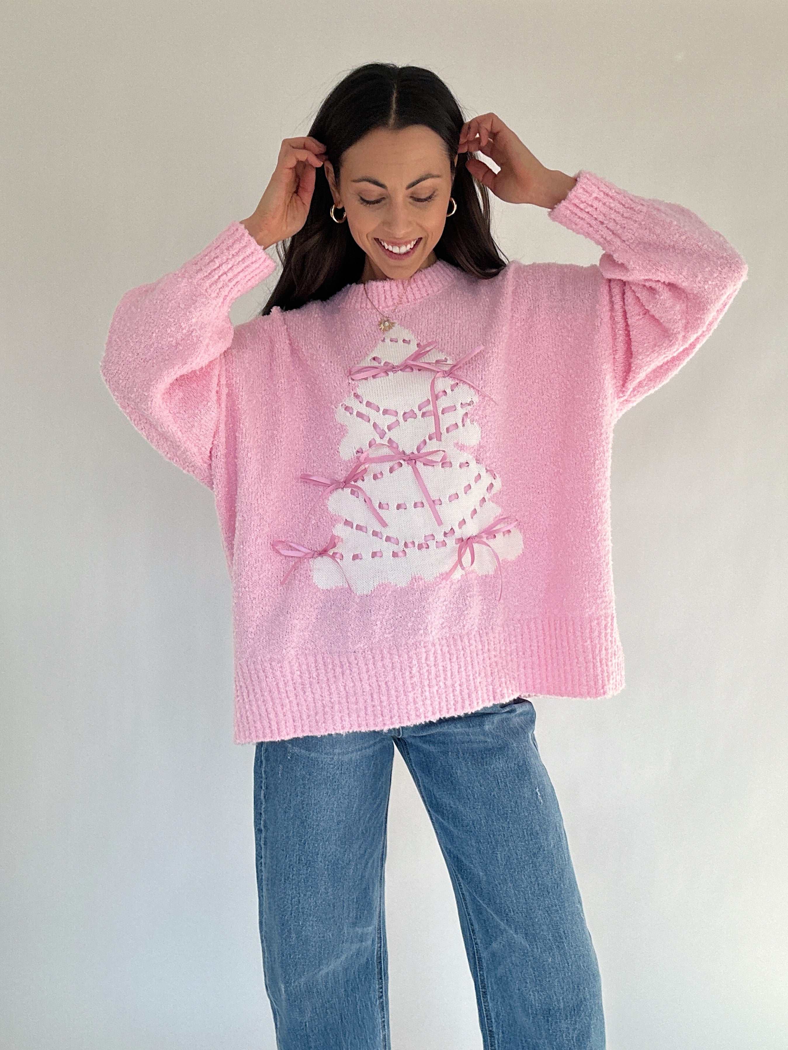 Person wearing a pink sweater with a tree design and blue jeans against a plain background