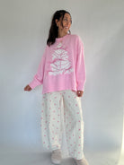 Woman wearing a pink sweater with a white tree design and white polka dot pants against a plain background