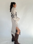 Woman wearing a long, patterned sweater dress with a side slit on a plain background showing side view