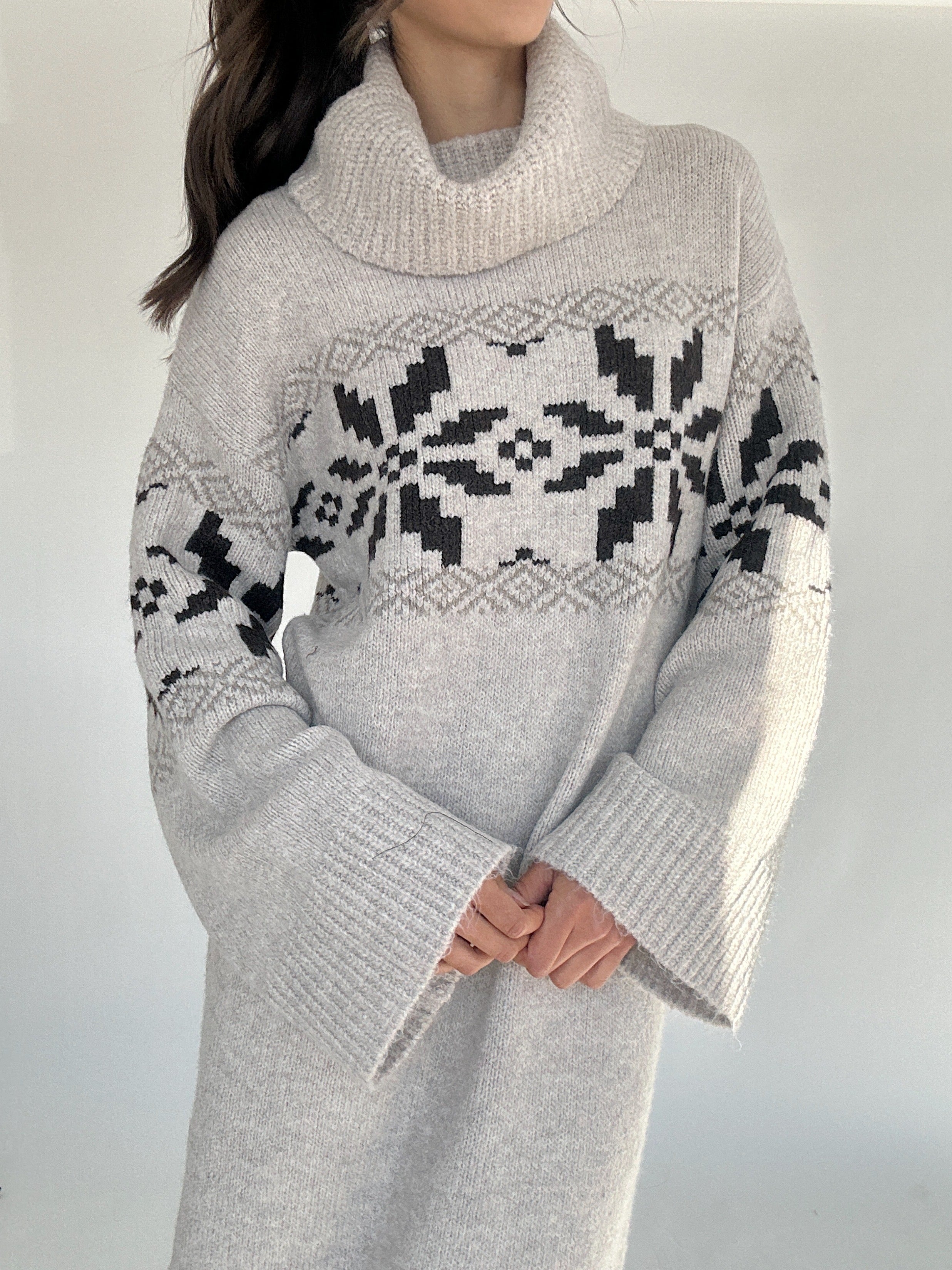 Person wearing a gray sweater dress with a black and white winter pattern and cowl neckline on a plain background showing top detail