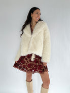Woman wearing a white faux fur jacket over a red patterned dress on a plain background