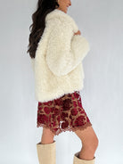 Person wearing a fluffy white jacket and patterned skirt on a plain background