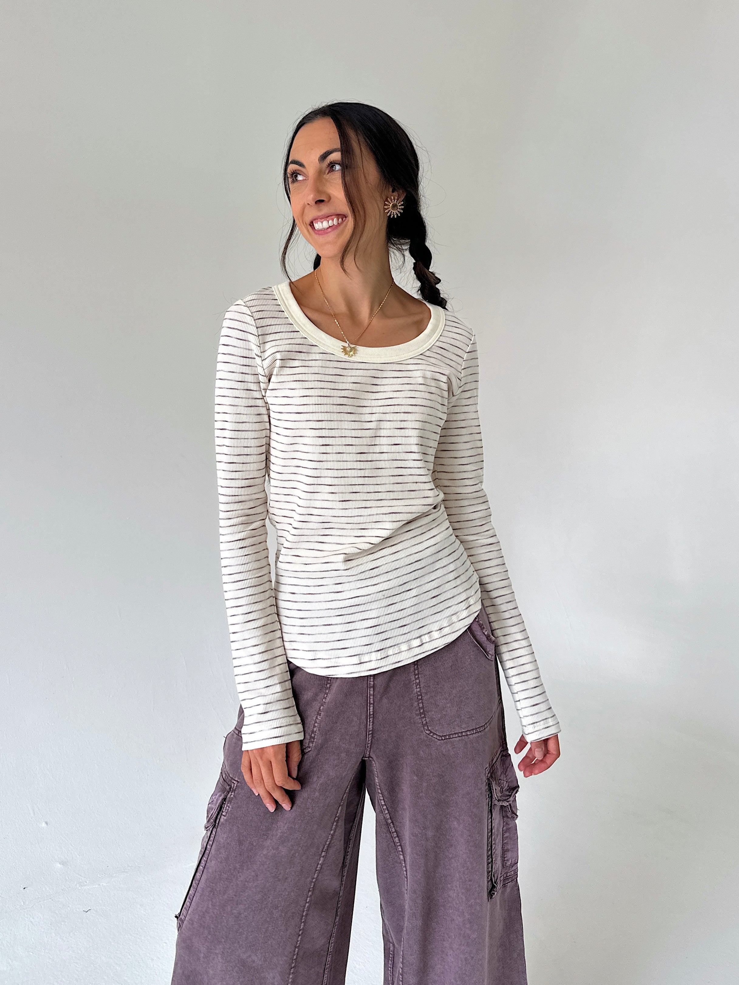 A woman modeling a cream-colored long-sleeve top with thin dark horizontal stripes, paired with wide-leg mauve cargo pants, against a plain light gray studio background.