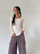 A woman with dark hair in two braids stands against a plain light gray wall, wearing a cream long-sleeve top with thin dark horizontal stripes and wide-leg mauve cargo pants.