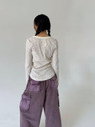A person shown from the back wearing a cream long-sleeve top with thin dark horizontal stripes and loose mauve-purple cargo pants, standing against a plain light gray wall.