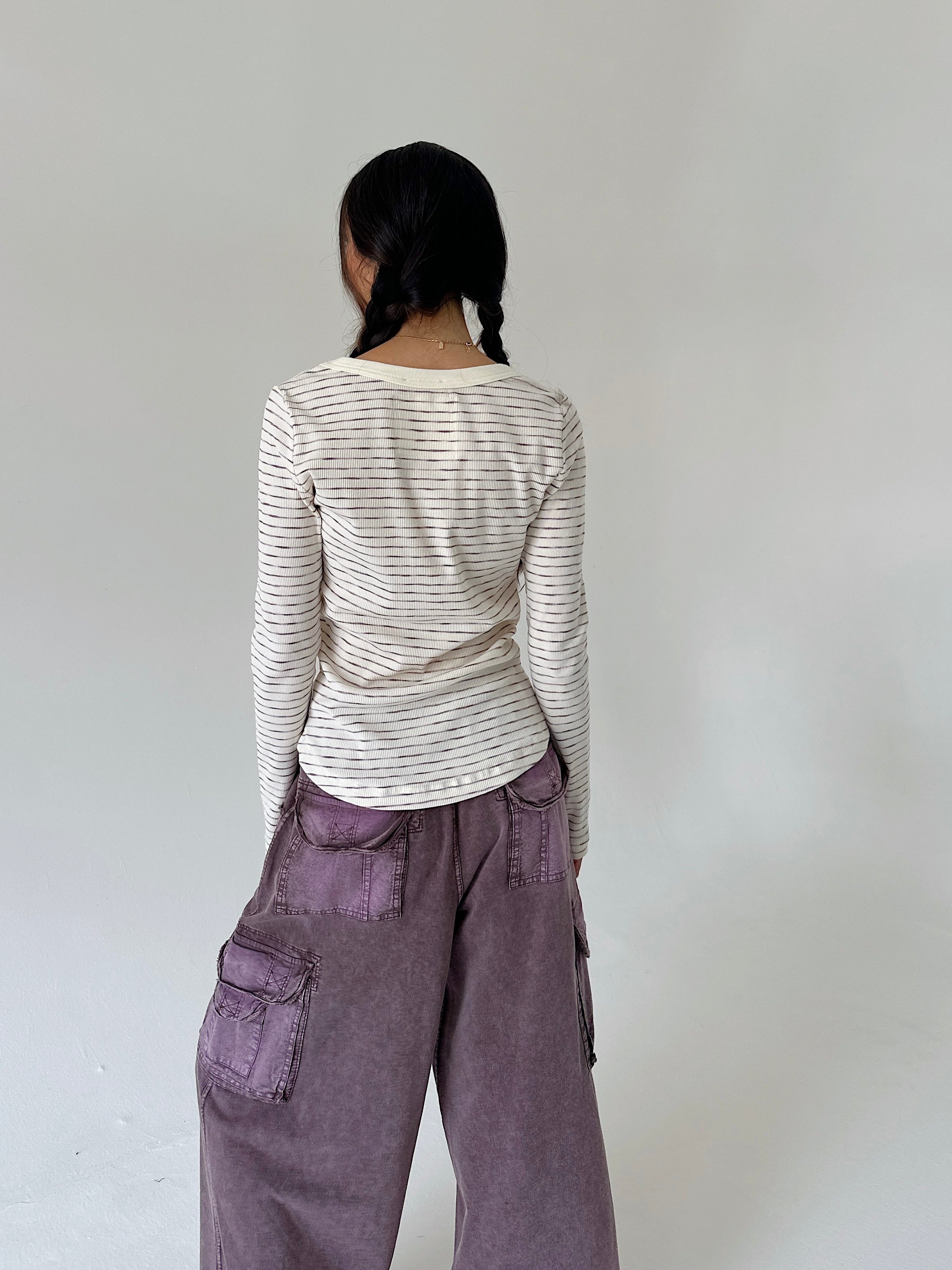 A person shown from the back wearing a cream long-sleeve top with thin dark horizontal stripes and loose mauve-purple cargo pants, standing against a plain light gray wall.