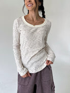 A woman wearing a cream-colored long-sleeve ribbed top with thin dark horizontal stripes and a scoop neckline, paired with dusty purple cargo pants, against a plain light gray background.
