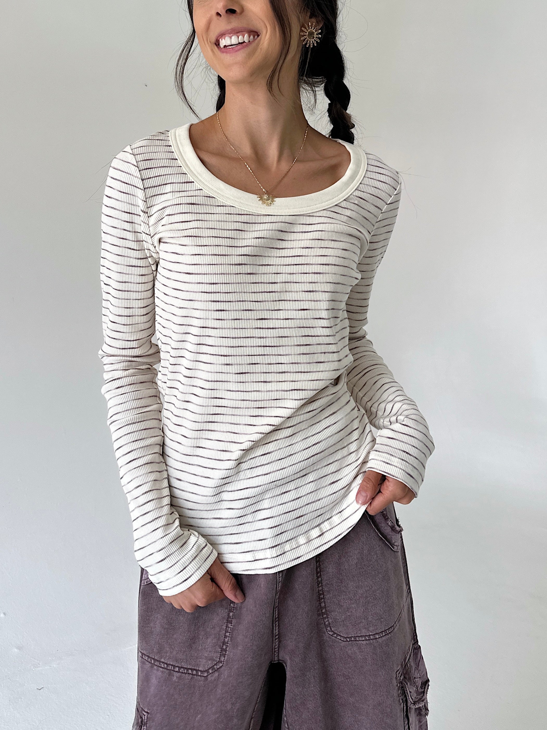 A woman wearing a cream-colored long-sleeve ribbed top with thin dark horizontal stripes and a scoop neckline, paired with dusty purple cargo pants, against a plain light gray background.