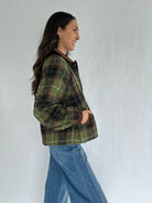 Woman wearing a green plaid jacket and blue jeans against a white background from side with hands in pockets