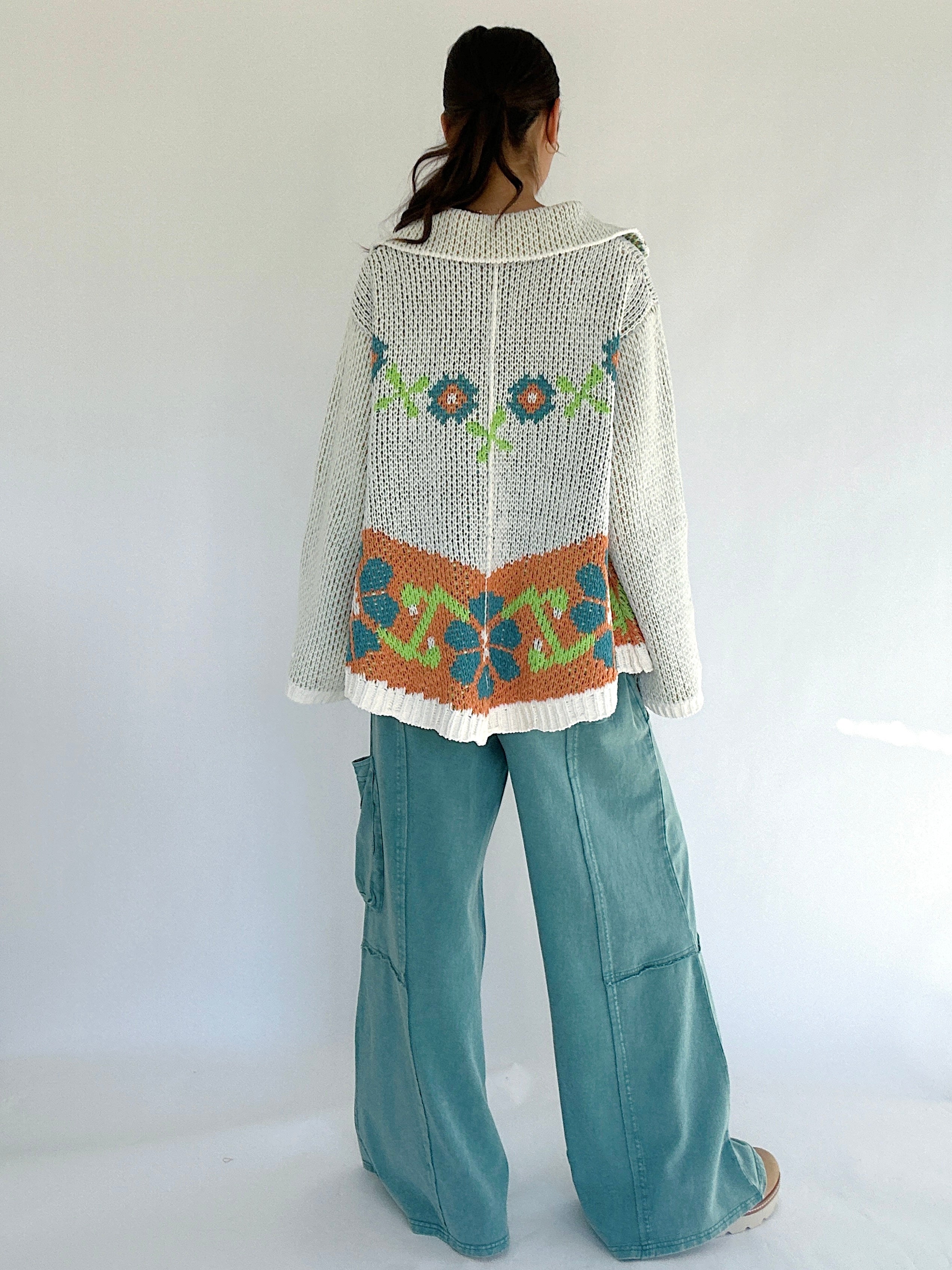 Person wearing a patterned sweater and teal pants on a white background showing back