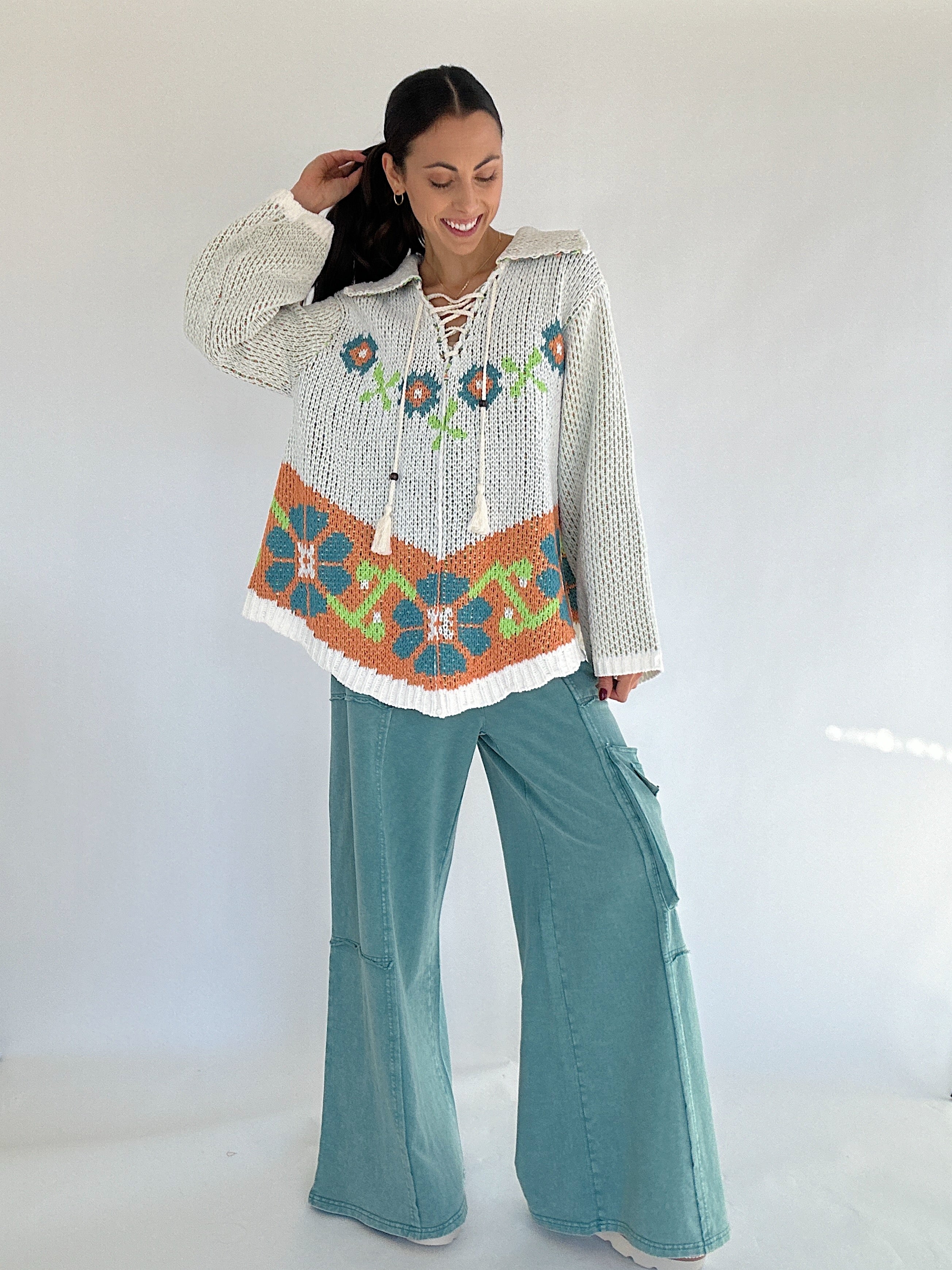 Woman wearing a patterned chenille sweater with orange and teal colors and a lace up neckline with teal pants against a white background