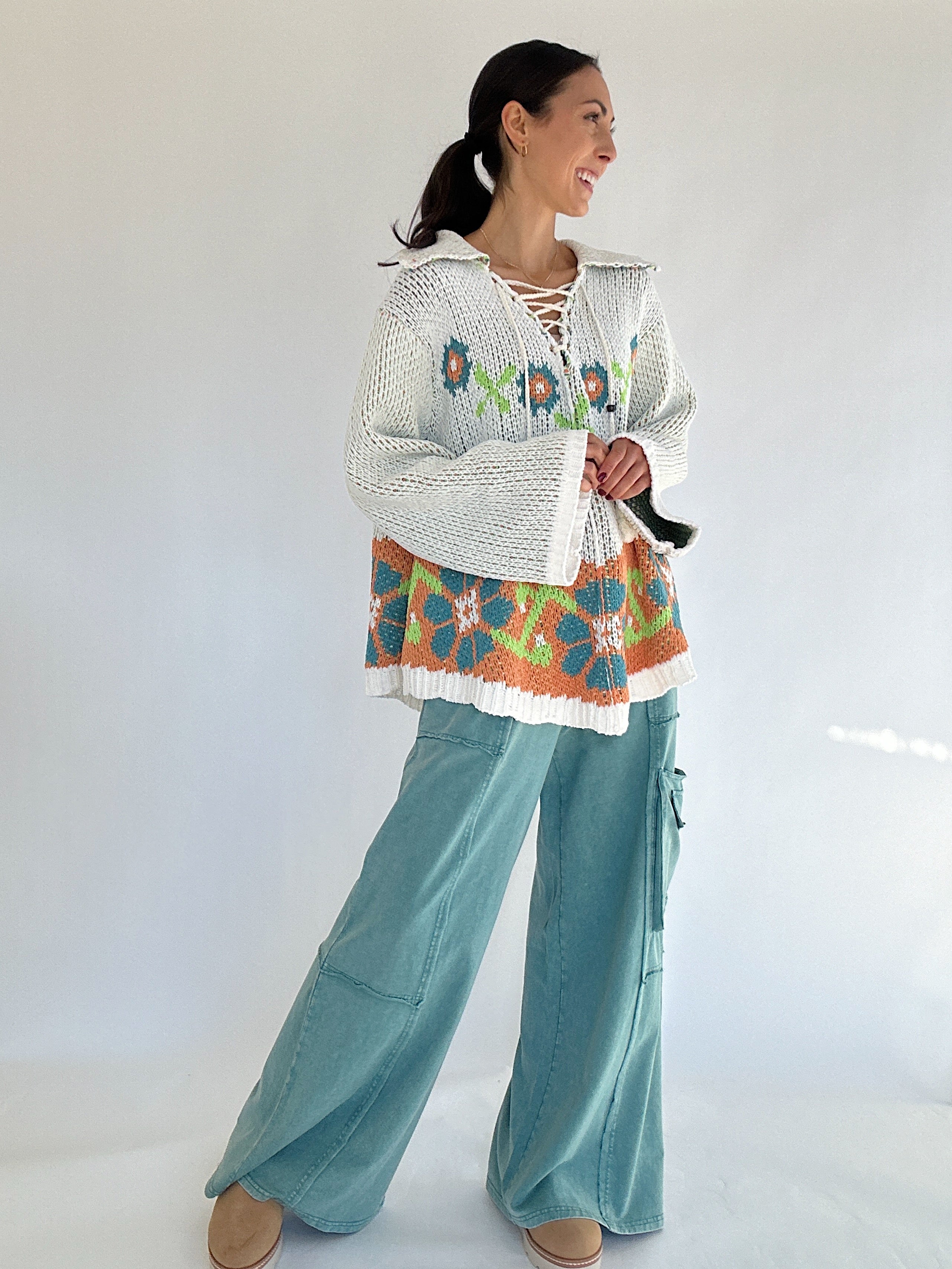 Woman wearing a colorful sweater with floral patterns and teal pants on a white background