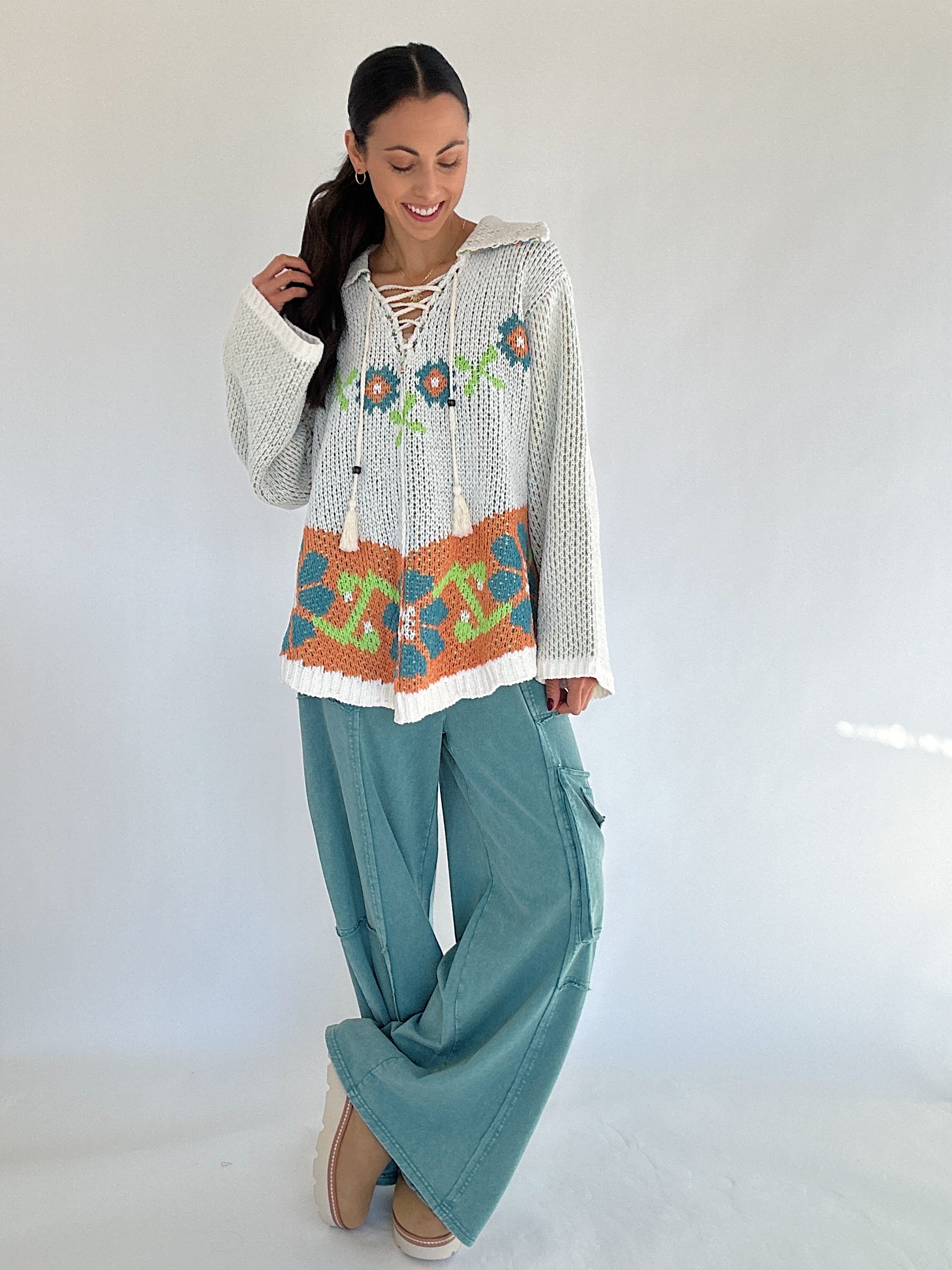 Woman wearing a patterned sweater and teal pants against a white background