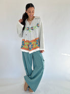 Woman wearing a patterned sweater and teal pants against a white background