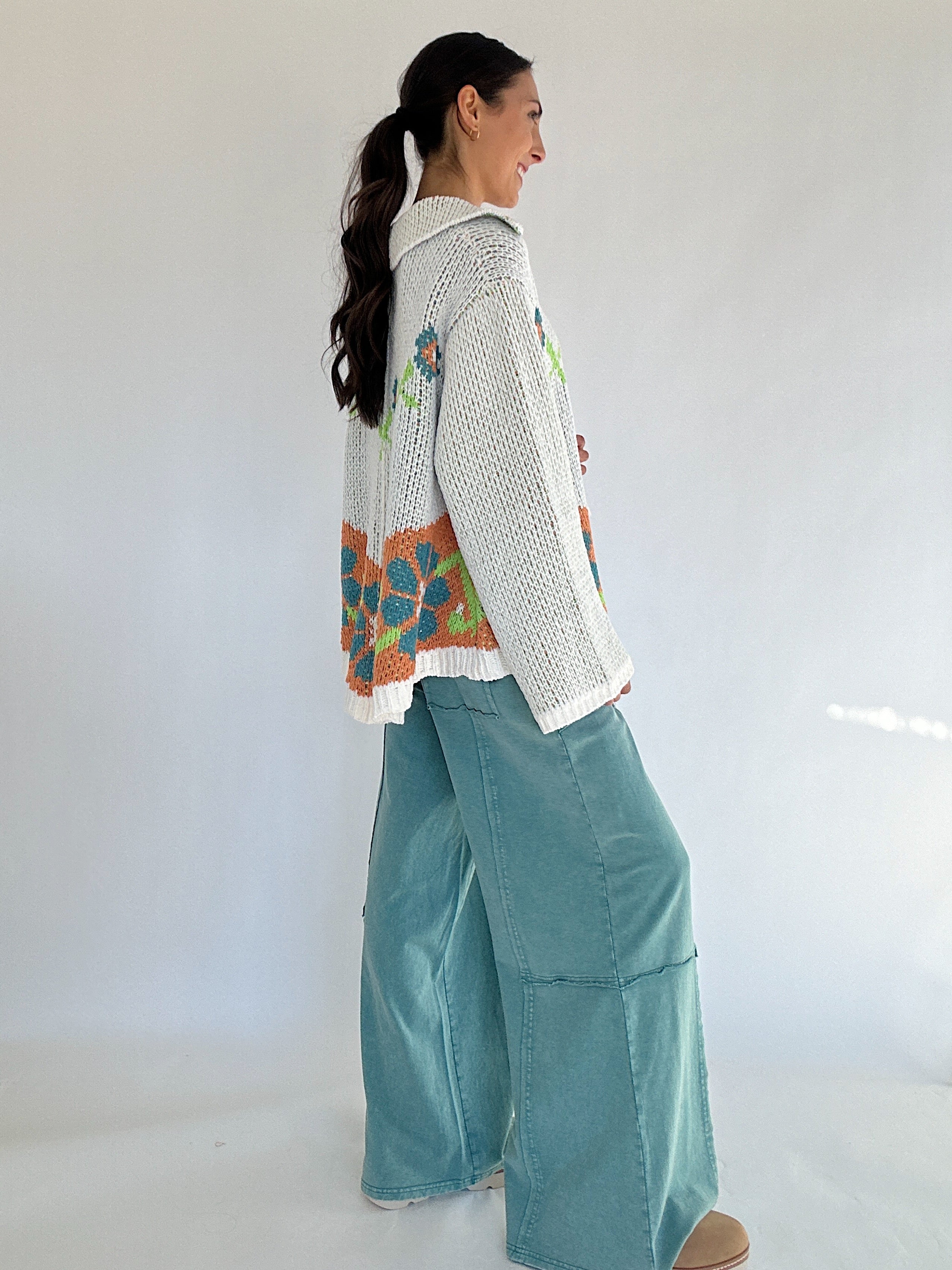 Woman wearing a colorful sweater and teal pants against a white background showing the side