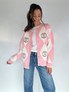 Woman wearing a pink and white argyle cardigan sweater with ornament embroidered designs, standing against a plain background.