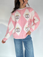 Person wearing a pink and white argyle and ornament patterned cardigan buttoned with blue jeans on a plain background