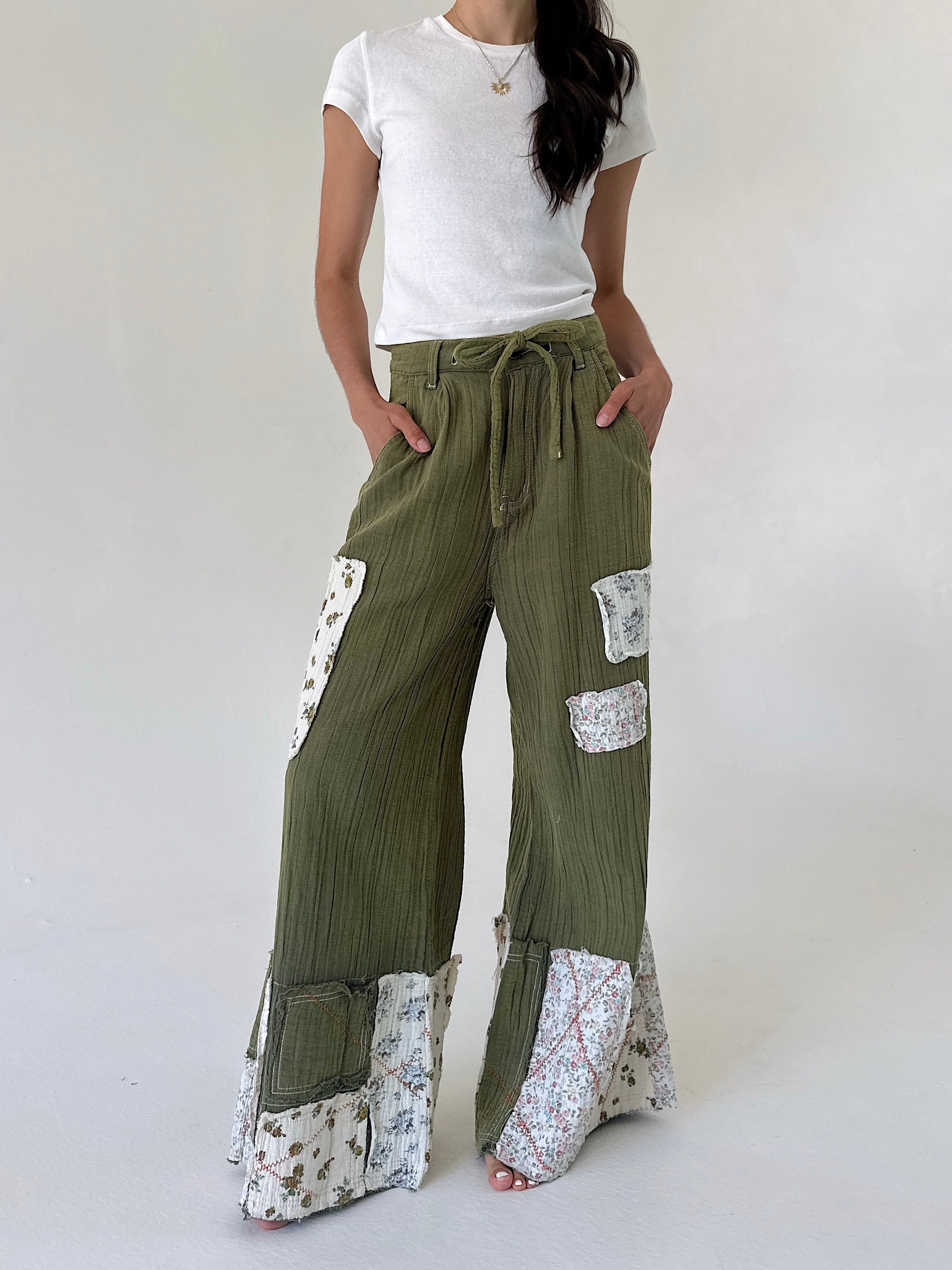 A model wearing olive green wide-leg patchwork pants with floral patterned inserts and a drawstring waist, paired with a white t-shirt against a plain light background.