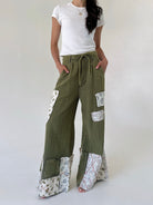 A model wearing olive green wide-leg patchwork pants with floral patterned inserts and a drawstring waist, paired with a white t-shirt against a plain light background.