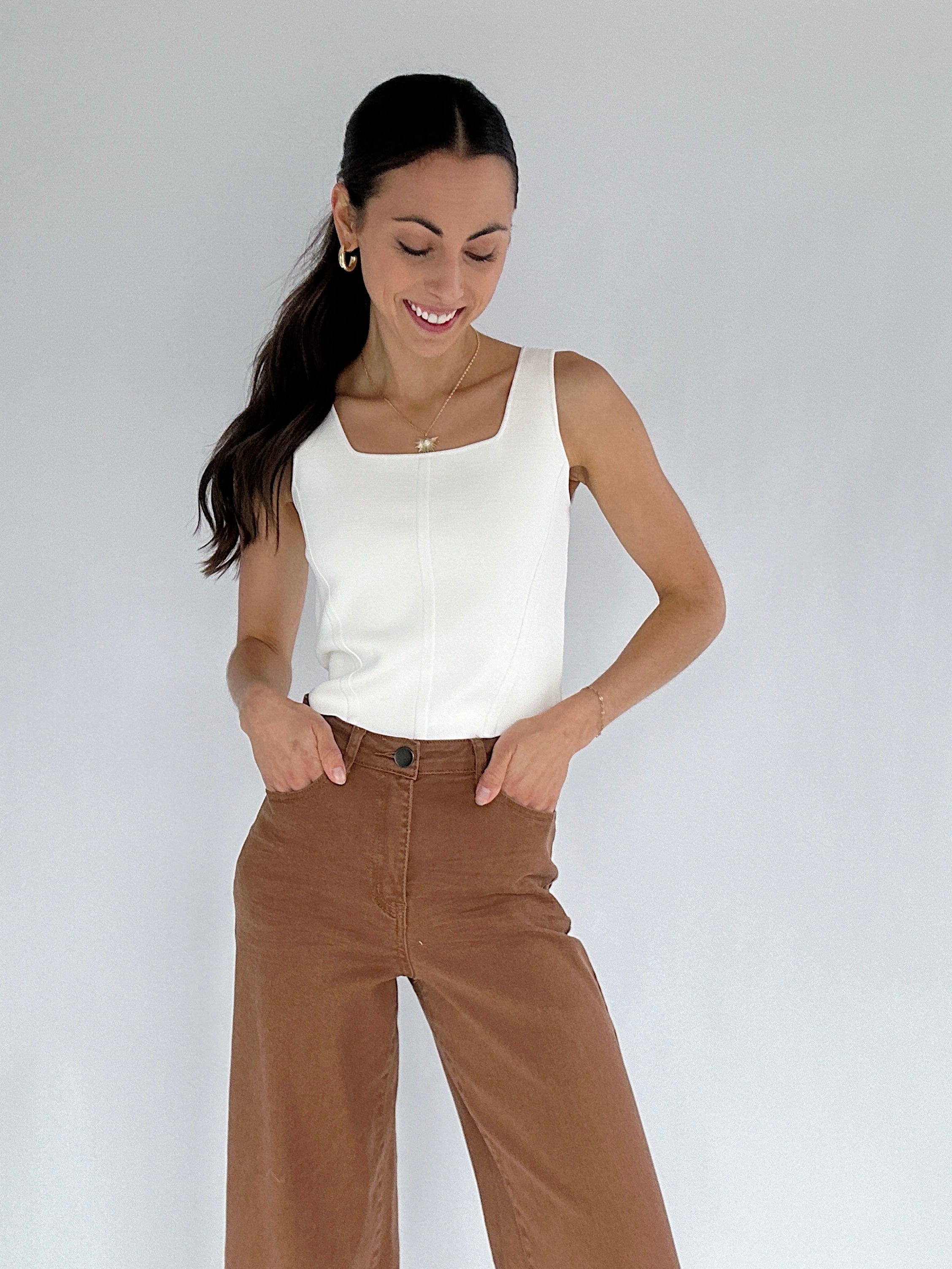 Woman wearing a white tank top and brown pants against a plain background
