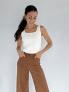 Woman wearing a white tank top and brown pants against a plain background