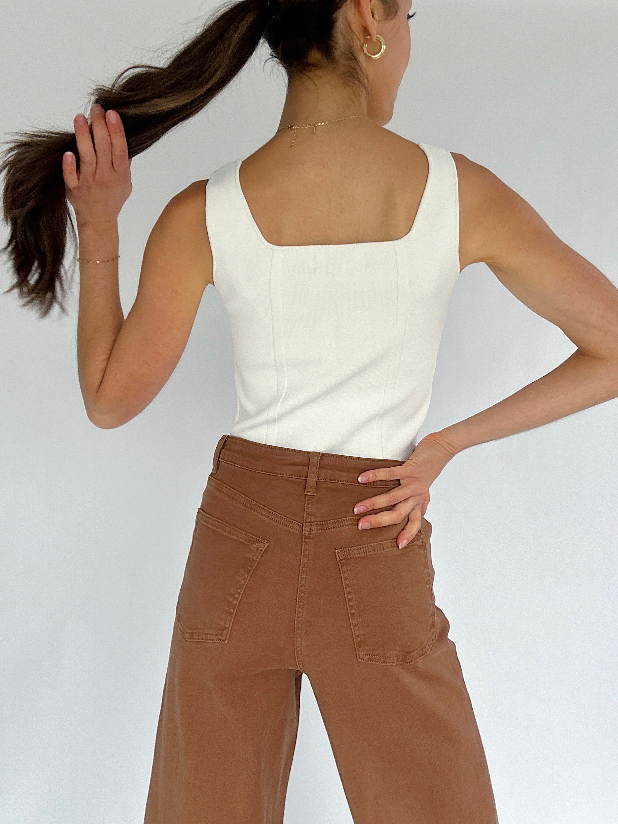 Person wearing a white sleeveless top and brown pants against a plain background showing back