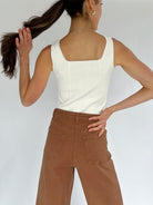 Person wearing a white sleeveless top and brown pants against a plain background showing back