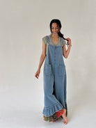 Woman wearing a denim overalls with a ruffle hem and legs crossed against a plain background