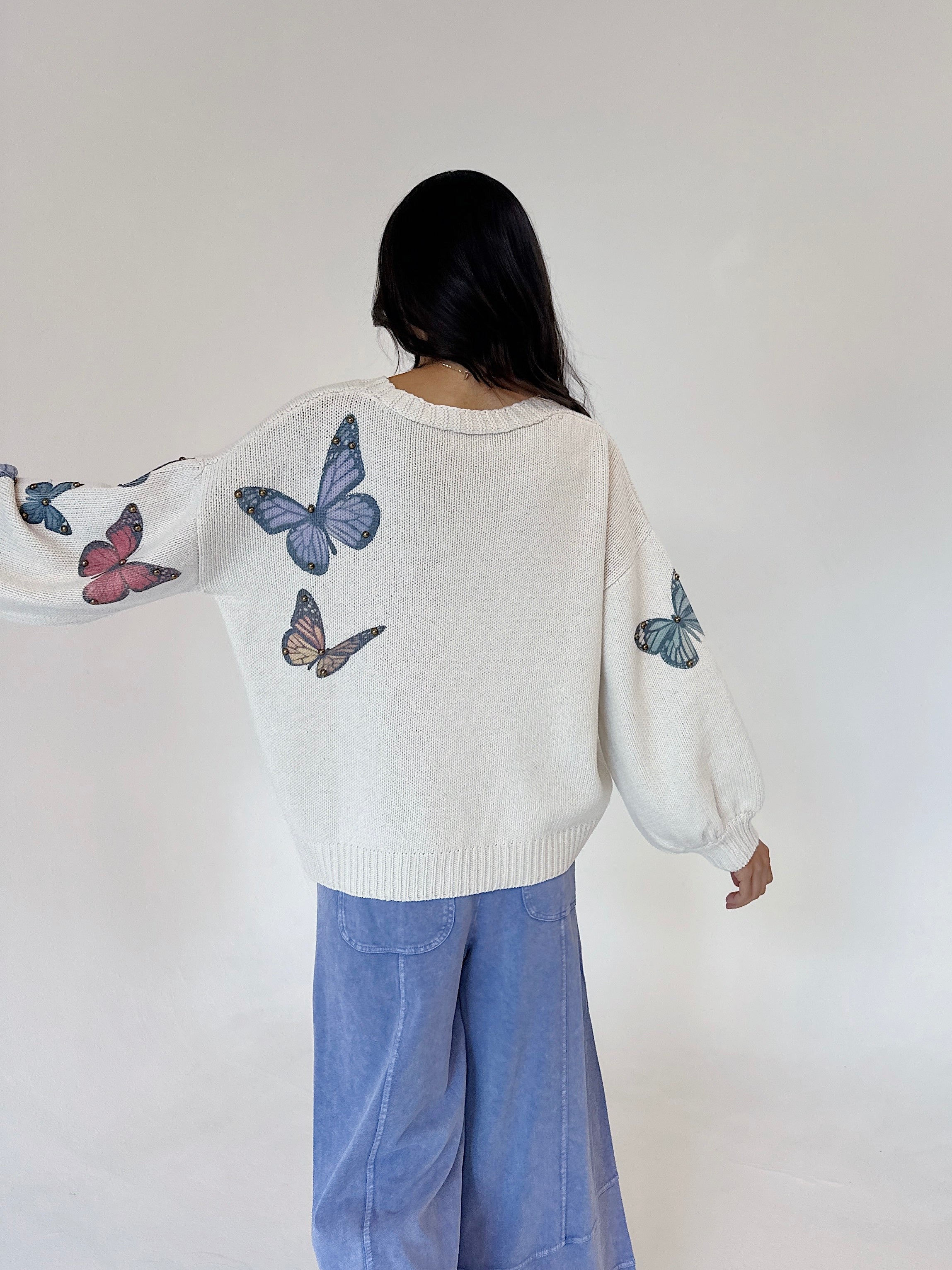 A person shown from behind wearing a cream knit sweater with blue, pink, and brown butterfly prints on the sleeves and back, paired with light blue wide-leg pants, against a plain off-white background.