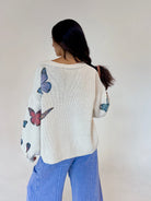 A woman viewed from behind wearing a cream-colored knit sweater with colorful butterfly prints on the back and sleeves, paired with light blue jeans, against a plain off-white wall.
