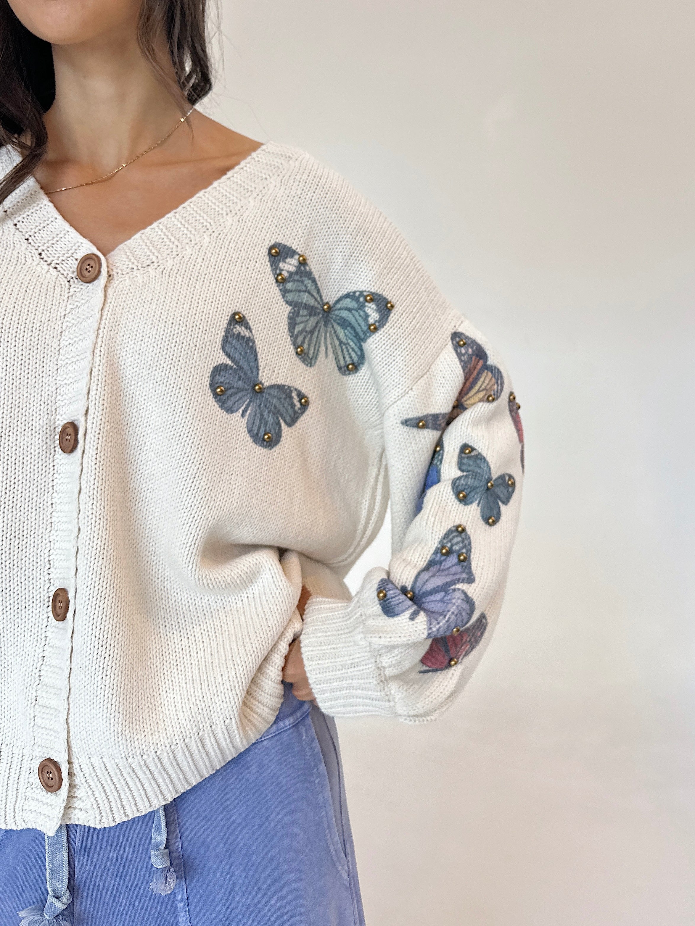 A close-up of a person wearing a cream knit cardigan with embroidered blue butterflies on the front and sleeves, paired with light blue jeans against a plain light background.