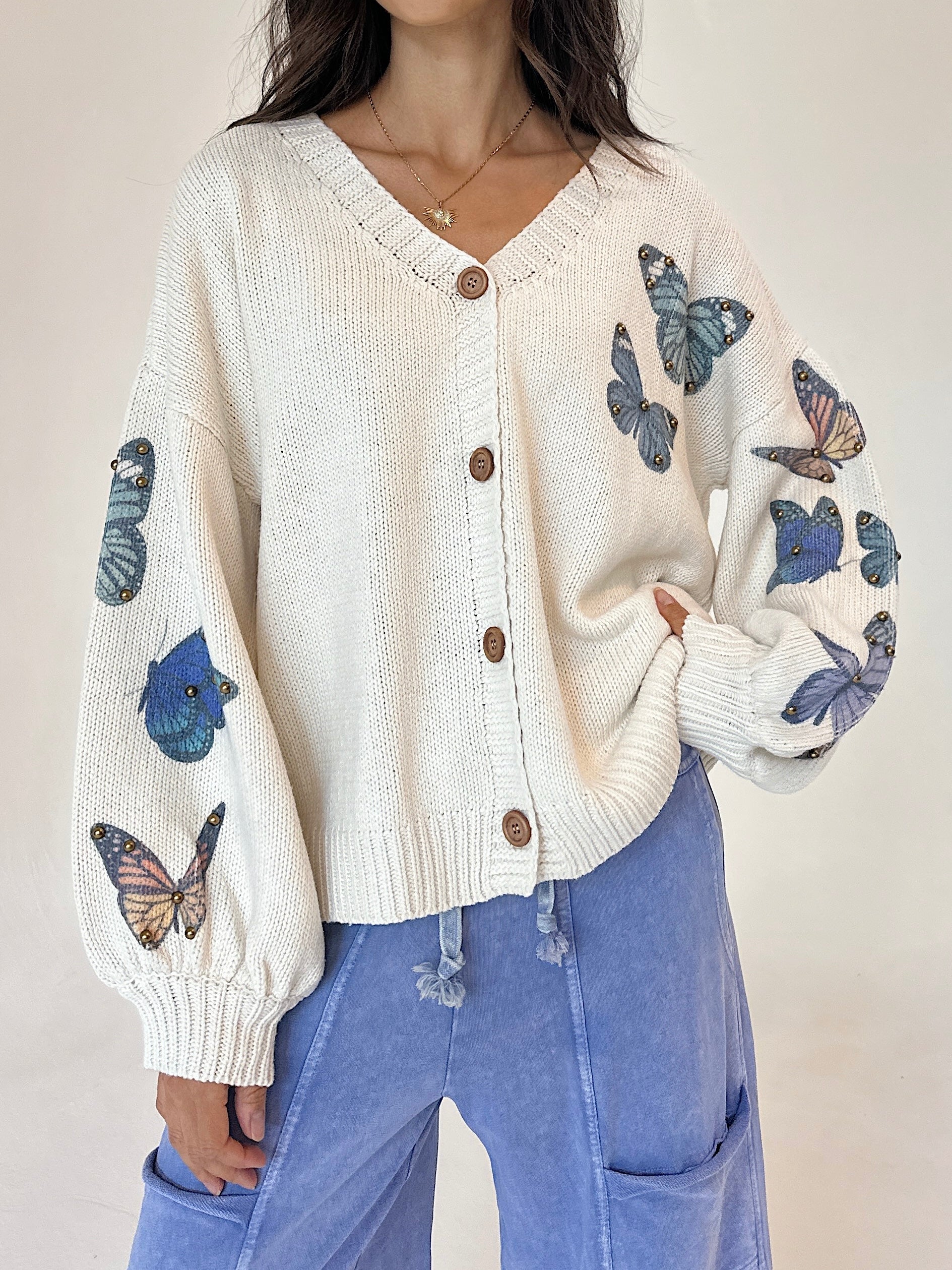 A woman wearing a cream-colored knit cardigan with blue and orange butterfly prints on the sleeves, paired with light blue pants, against a neutral beige background.
