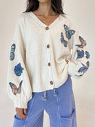 A woman wearing a cream-colored knit cardigan with blue and orange butterfly prints on the sleeves, paired with light blue pants, against a neutral beige background.