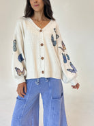 A woman wearing a cream-colored knit cardigan with brown buttons and blue butterfly prints on the sleeves, paired with light blue wide-leg pants, against a plain off-white background.
