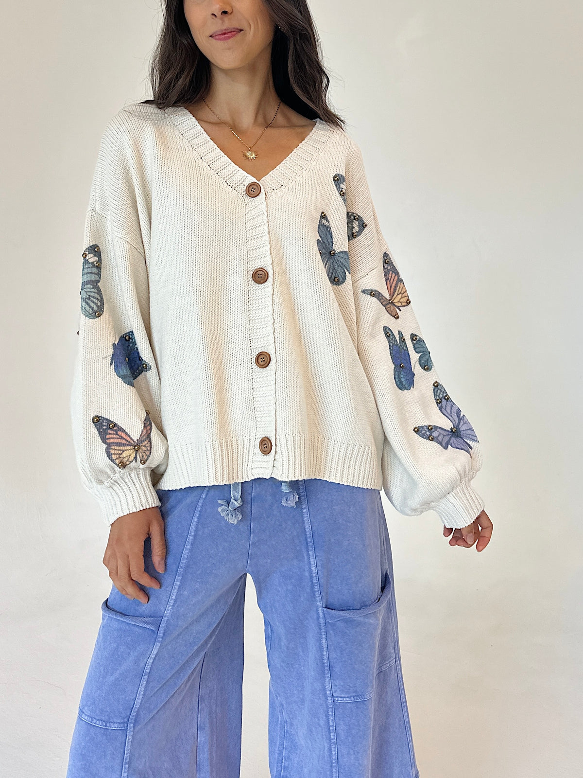 A woman wearing a cream-colored knit cardigan with brown buttons and blue butterfly prints on the sleeves, paired with light blue wide-leg pants, against a plain off-white background.