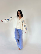 A woman modeling a cream-colored knit cardigan with blue butterfly embroidery on the sleeves, paired with light blue wide-leg pants, against a plain off-white studio background.