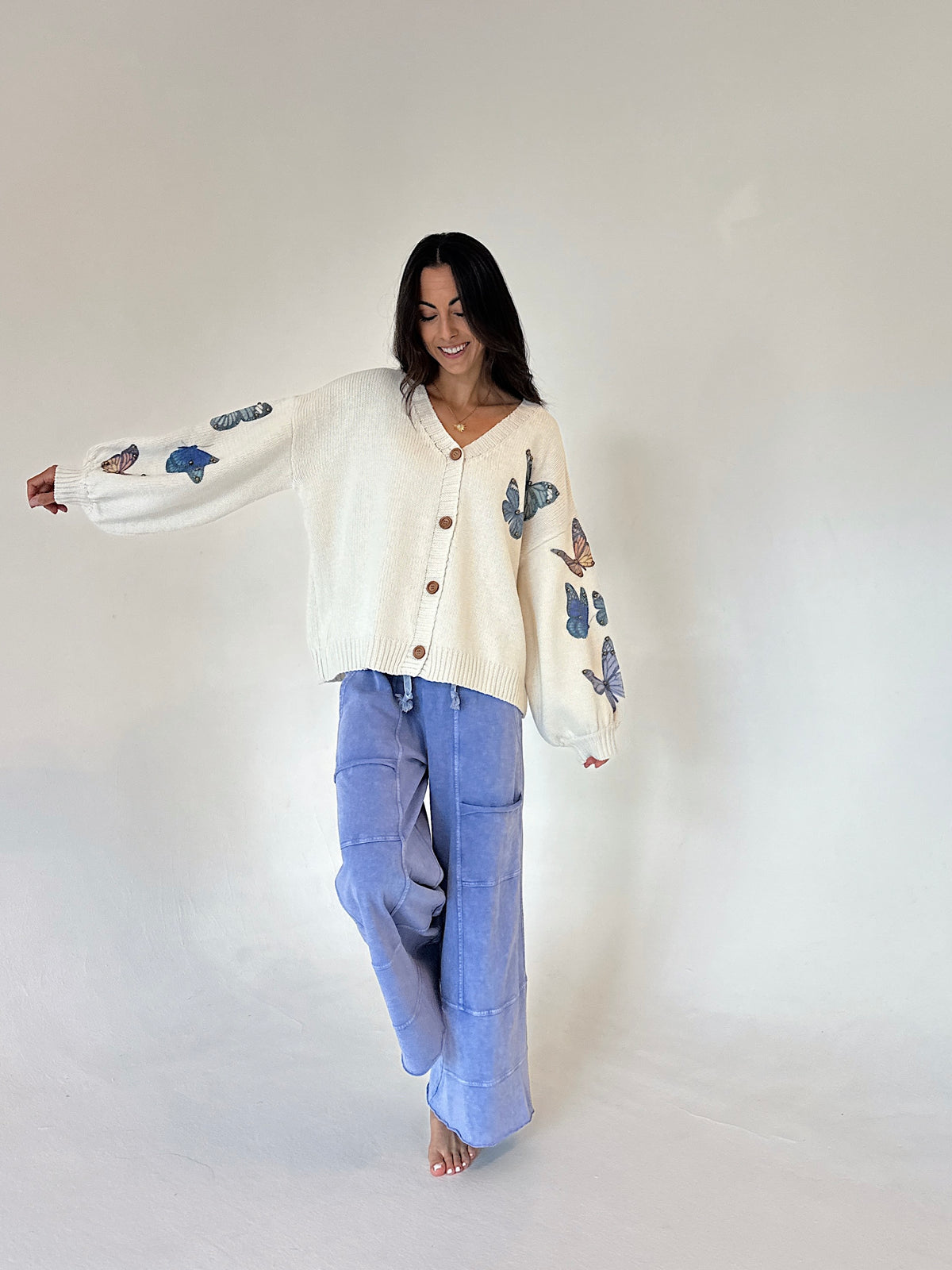 A woman modeling a cream-colored knit cardigan with blue butterfly embroidery on the sleeves, paired with light blue wide-leg pants, against a plain off-white studio background.