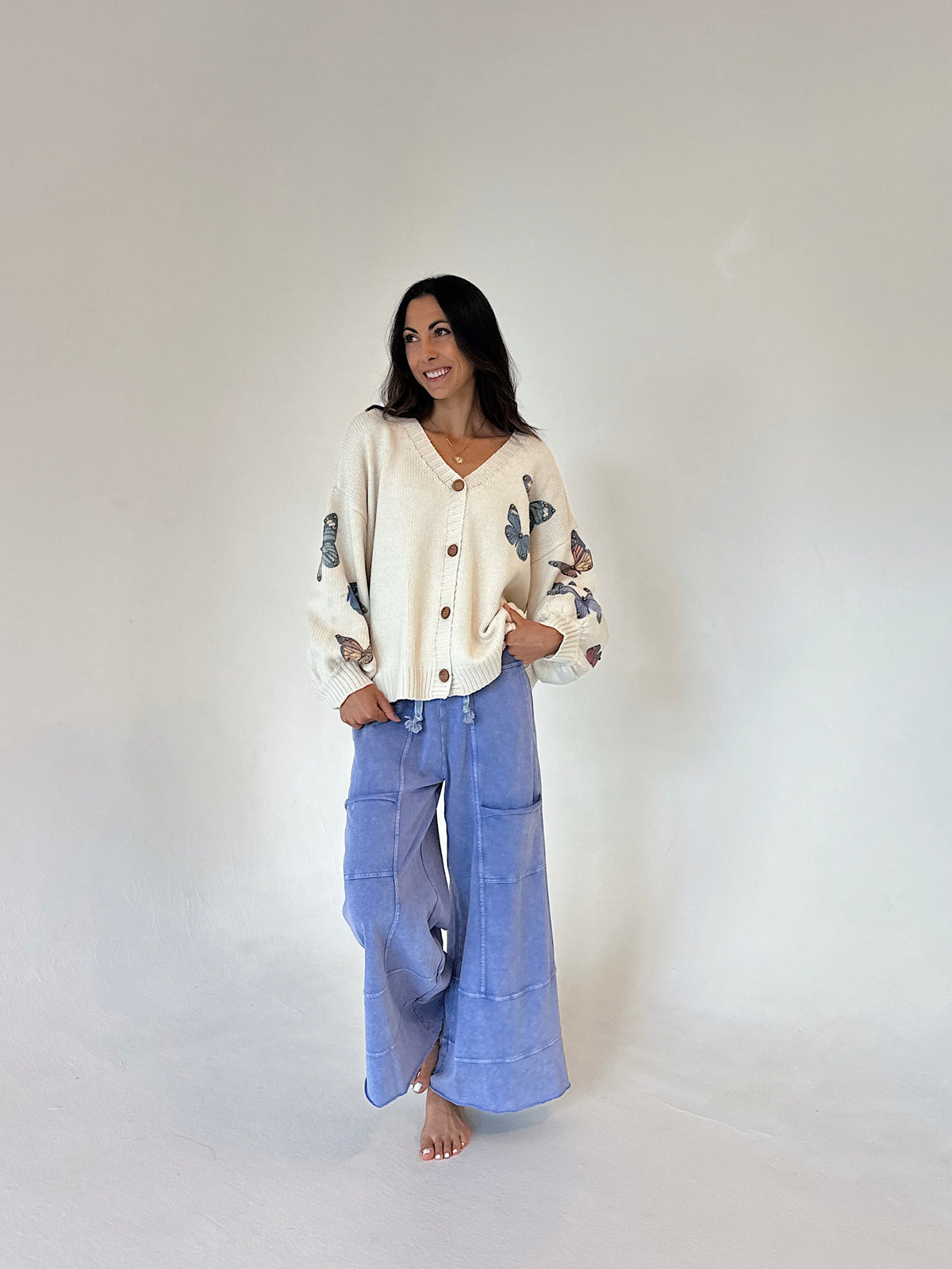A woman stands barefoot against a plain light gray studio background, wearing a cream cardigan with blue butterfly embroidery on the sleeves and blue wide-leg pants.