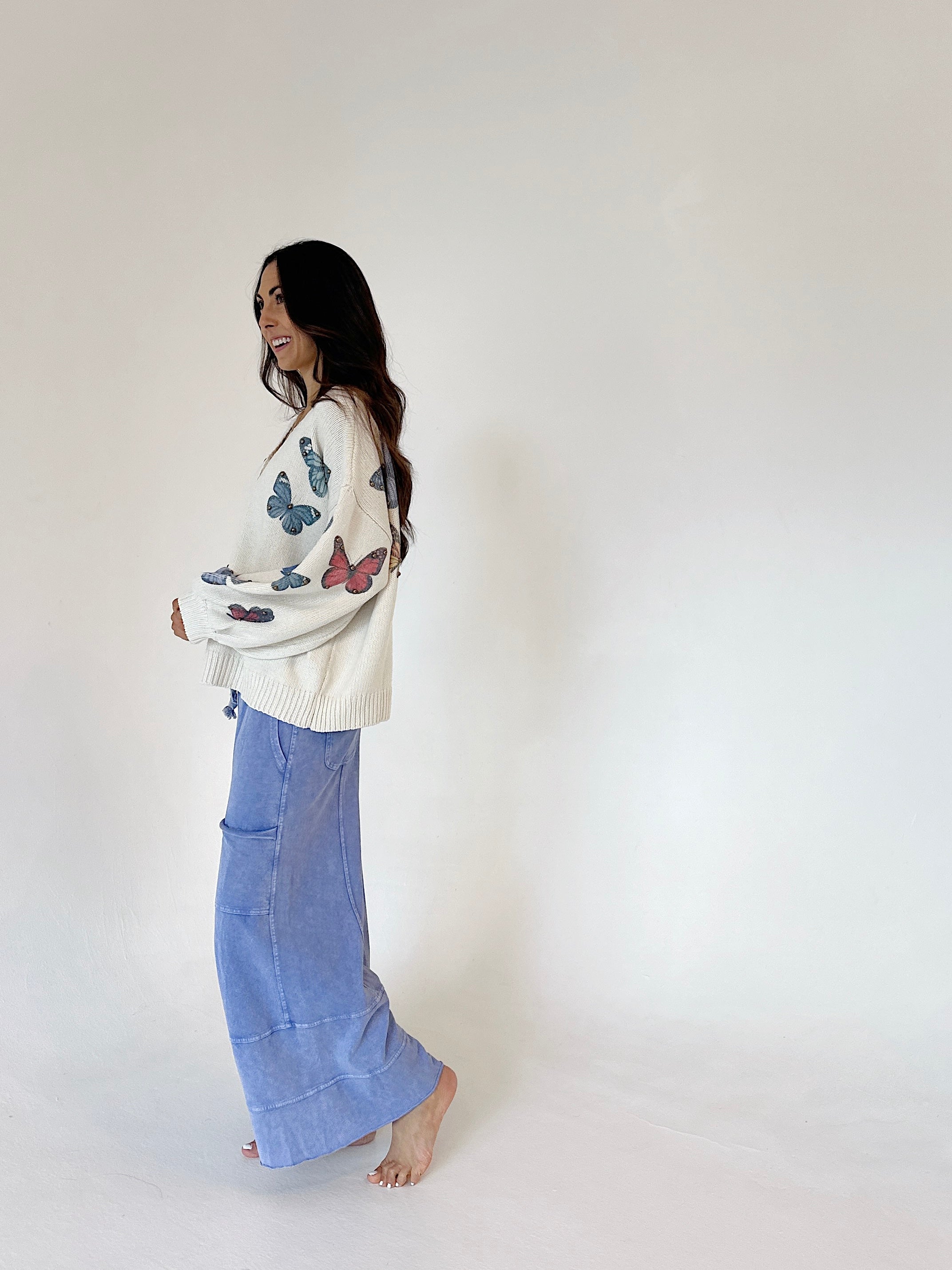 A woman stands barefoot against a plain white studio background, wearing a cream knit sweater with colorful butterfly prints and blue wide leg pants from side.