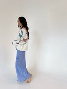 A woman stands barefoot against a plain white studio background, wearing a cream knit sweater with colorful butterfly prints and blue wide leg pants from side.