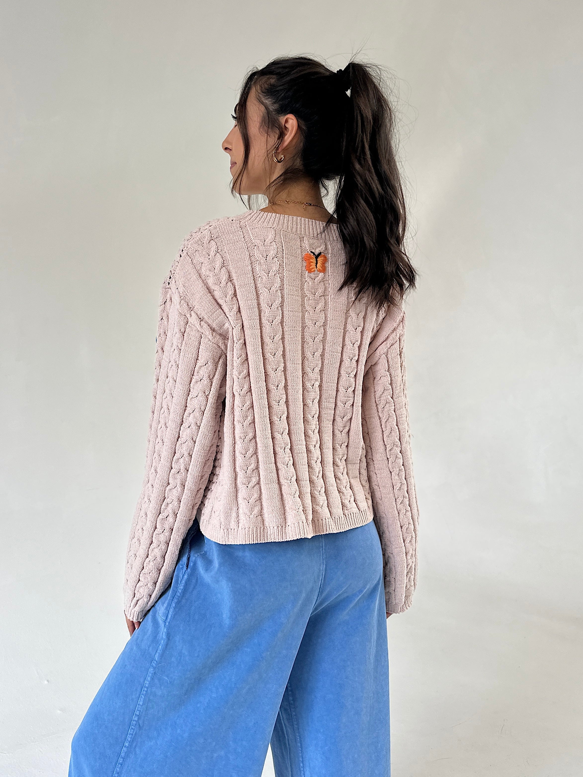 A woman shown from the back wearing a taupe cable-knit cropped sweater with a small orange butterfly graphic on the upper back, paired with wide-leg blue jeans, against a plain off-white background.