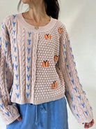 A woman wearing a taupe cable-knit sweater with blue patterns and orange butterfly appliqués, paired with blue pants against a plain white background.