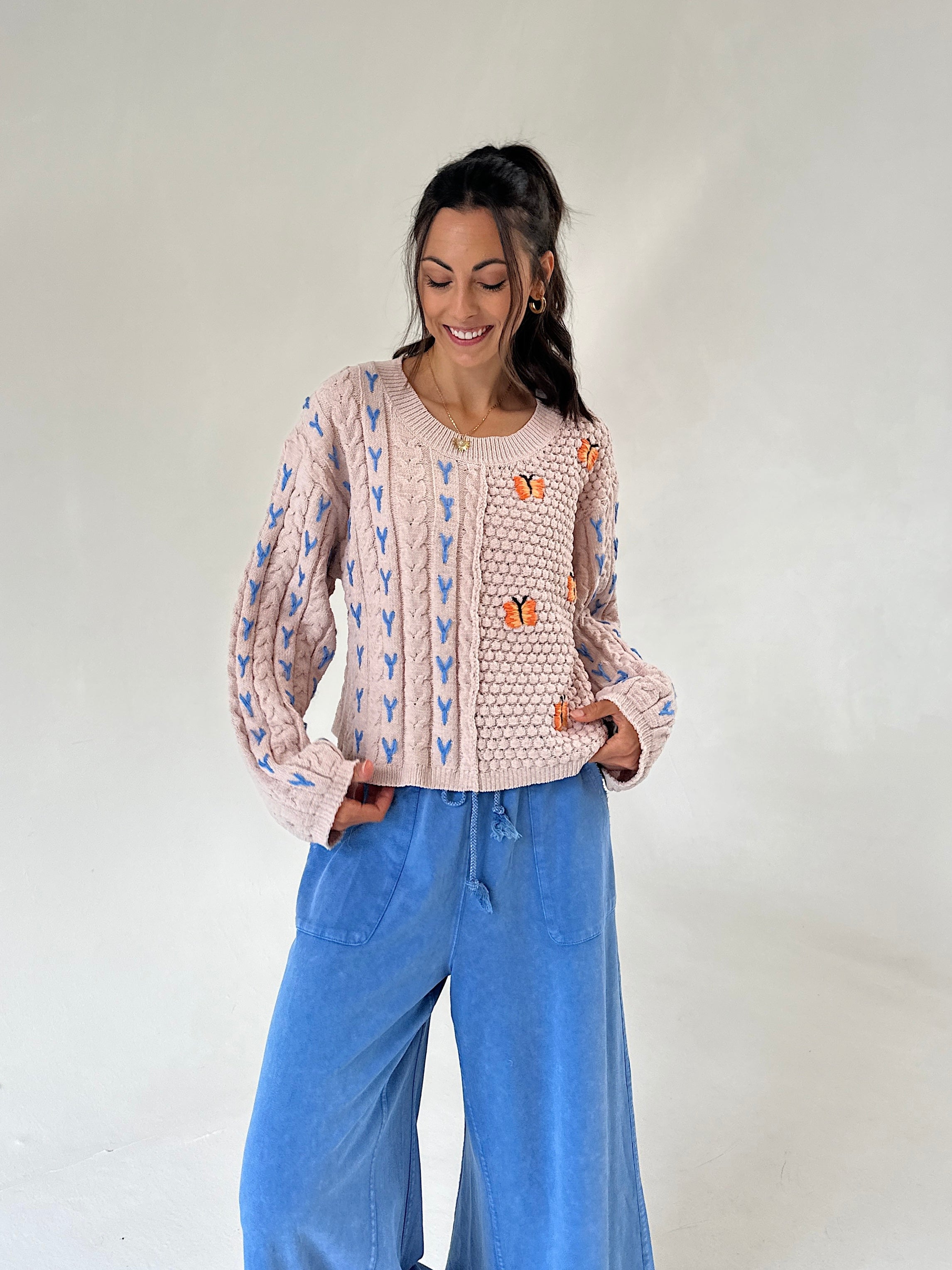 A woman modeling a taupe cable-knit sweater with blue and orange embroidered details, paired with wide-leg blue jeans, against a plain light gray studio background.