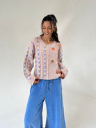 A woman modeling a taupe cable-knit sweater with blue and orange embroidered details, paired with wide-leg blue jeans, against a plain light gray studio background.