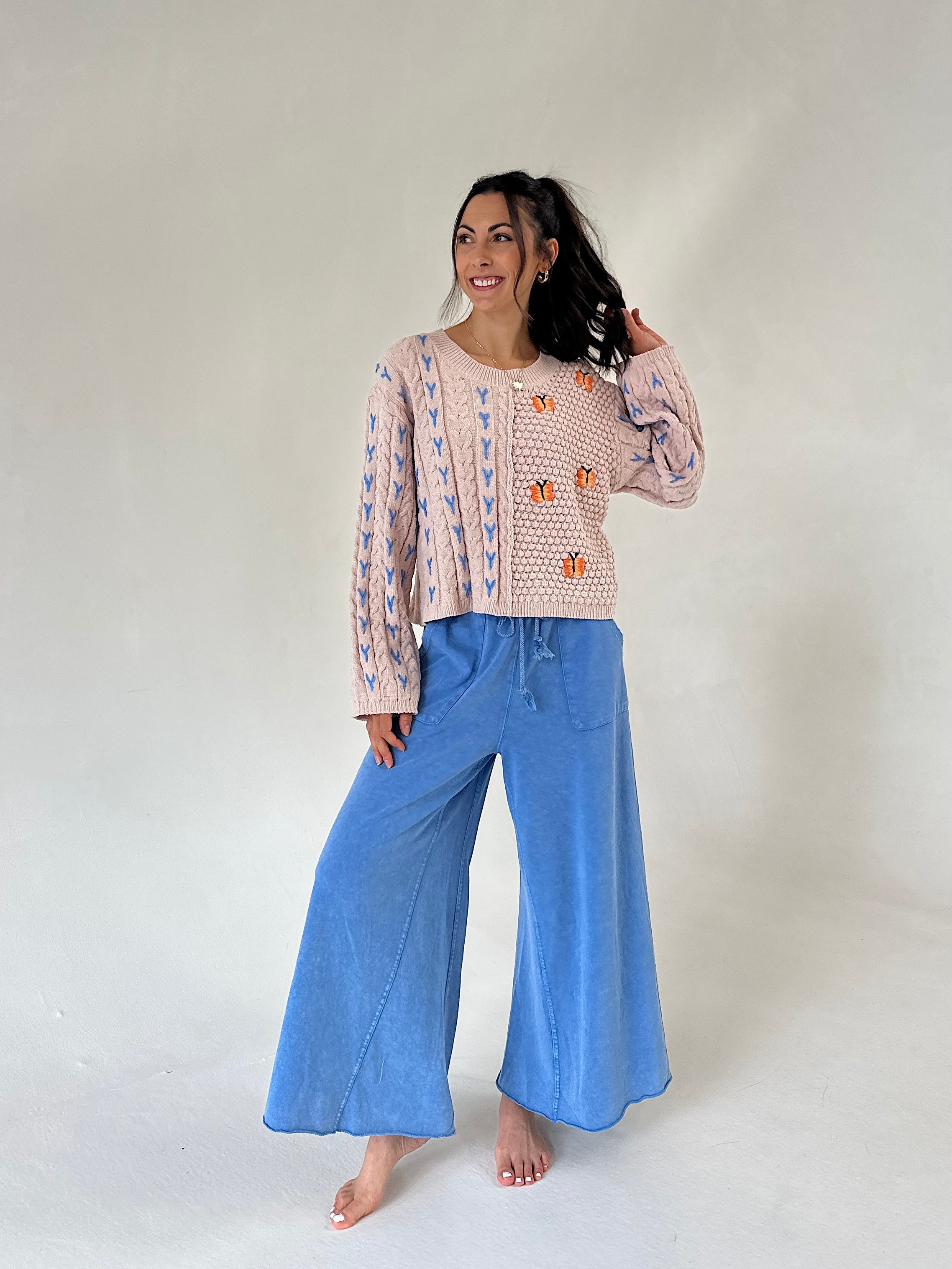 A woman modeling a taupe knit sweater with blue and orange patterns, paired with wide-leg blue pants, standing against a plain light gray studio background.